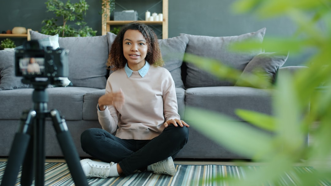 Happy Afro-American girl blogger is recording video for vlog showing thumbs-up hand gesture sitting on floor at home using camera on tripod.